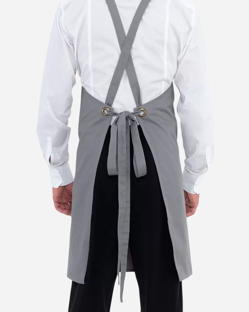 Grembiule unisex grigio in poliestere e cotone con pettorina, tasca centrale e allacciatura con bretelle regolabili tramite anello in ottone.